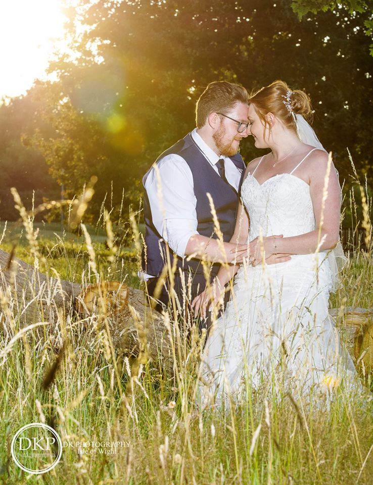 Couple in wedding attire embrace outdoors in front of a bright sun. Tall grass in foreground.