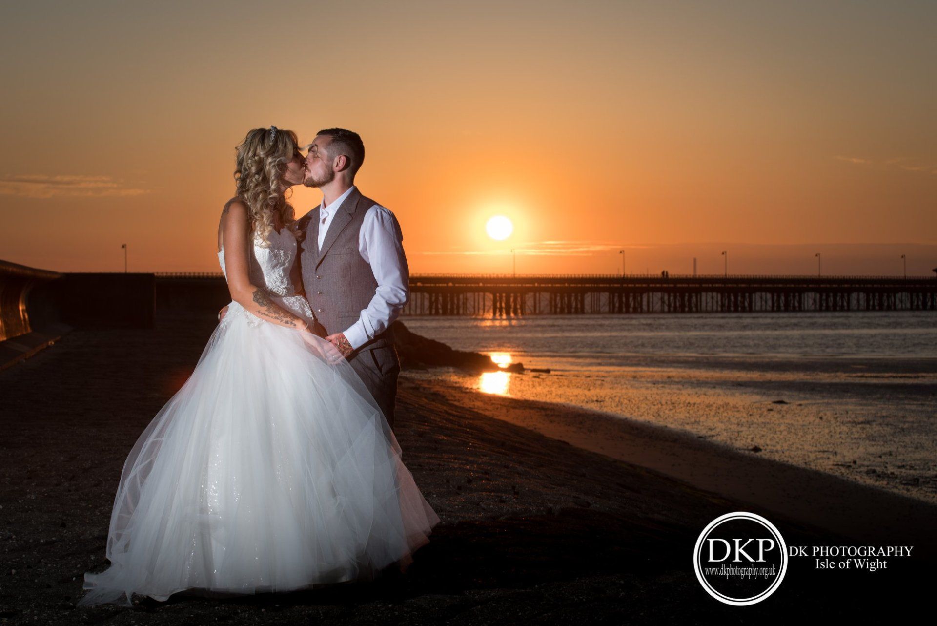 Bride and groom kissing at sunset on a beach, pier in the background.
