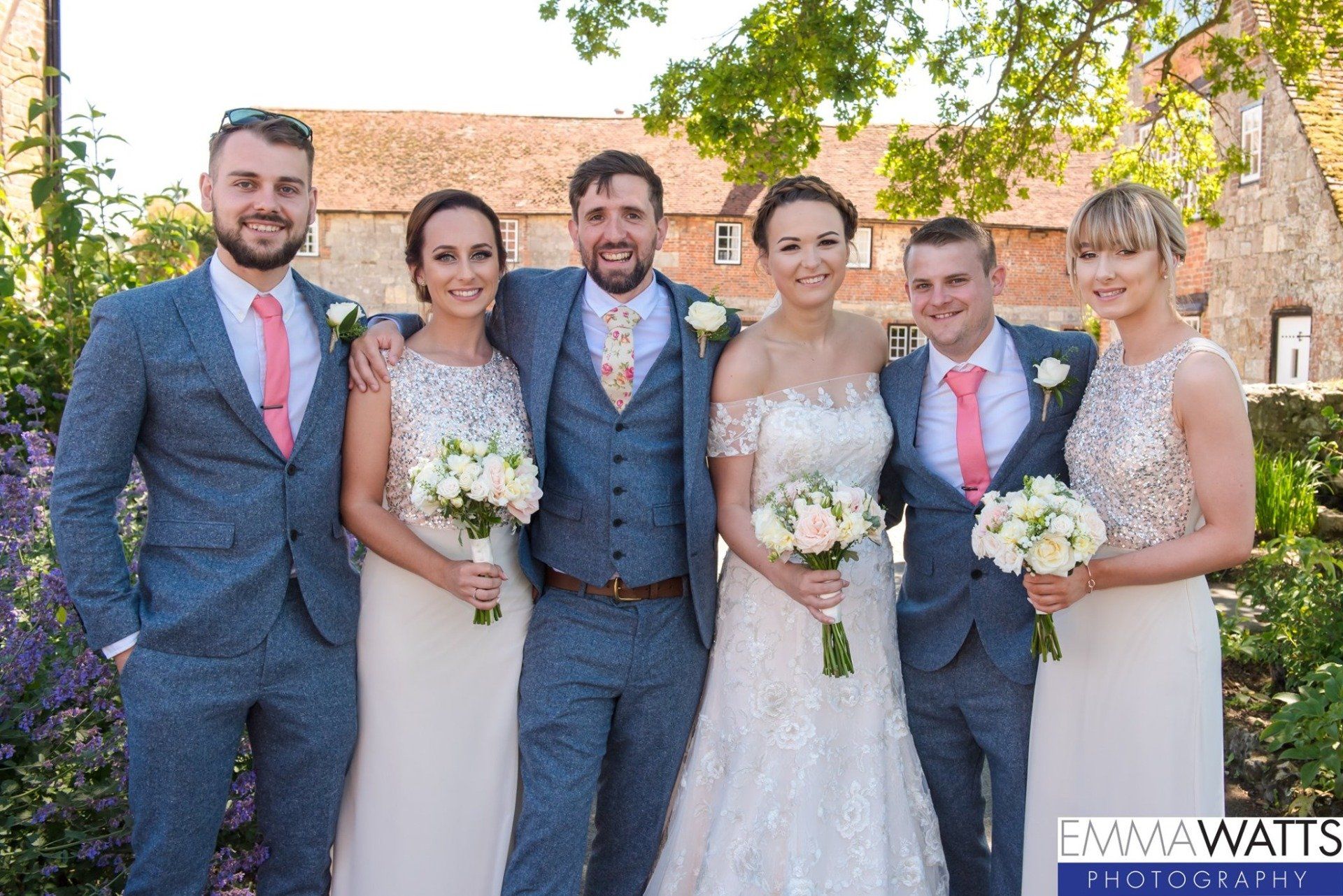 Wedding group: six people pose outdoors. Groom in blue suit and floral tie, bridesmaids in light dresses, pink ties.