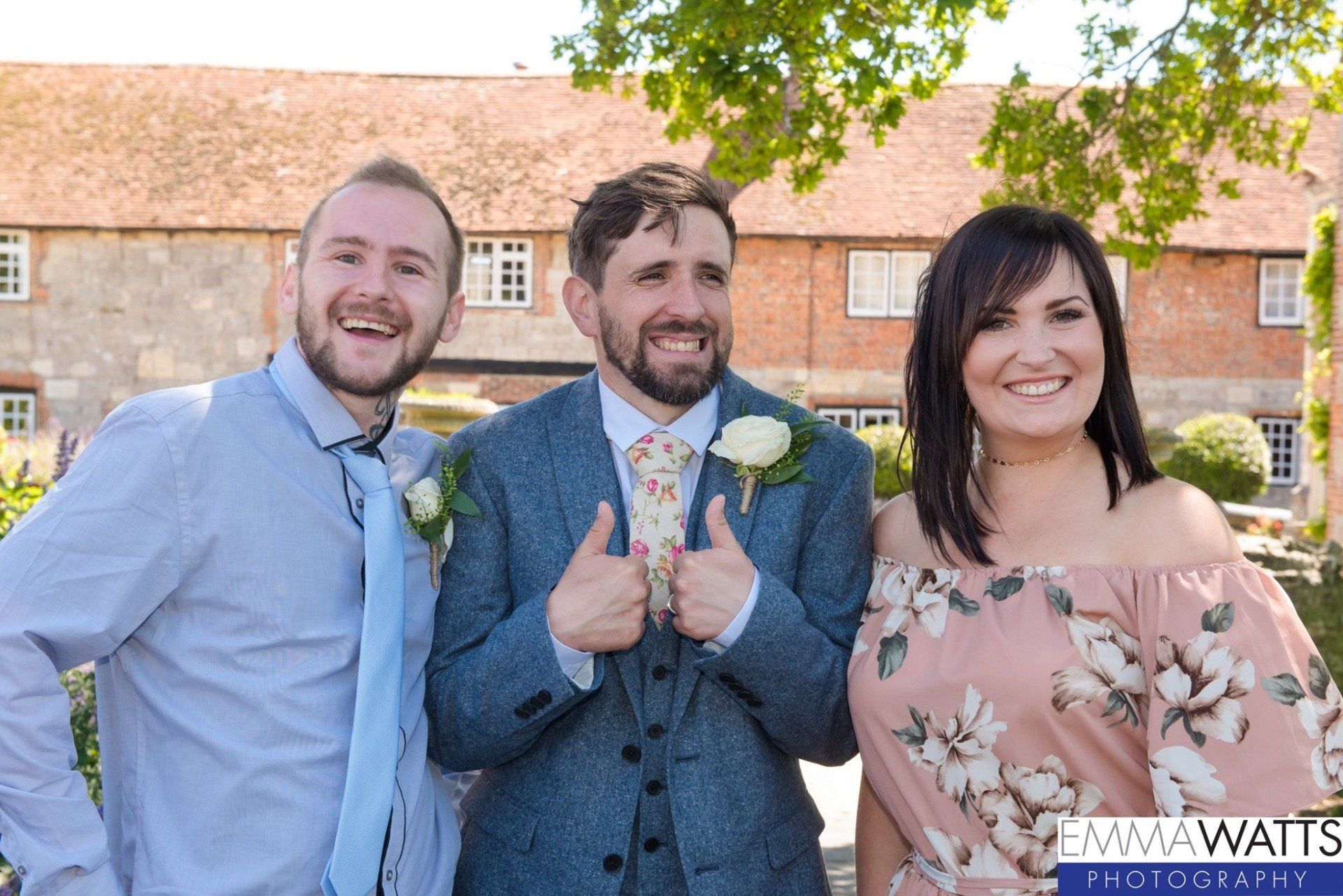 Three people smiling outside a brick building. Man in center gives thumbs up, wearing a suit.