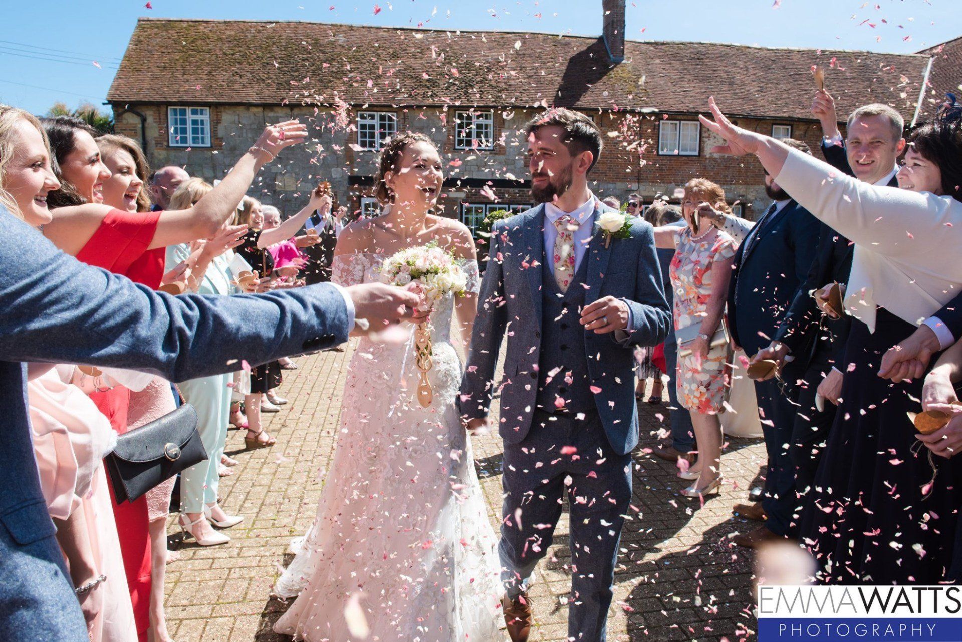 Bride and groom walk through confetti, smiling, flanked by guests, near a brick building.