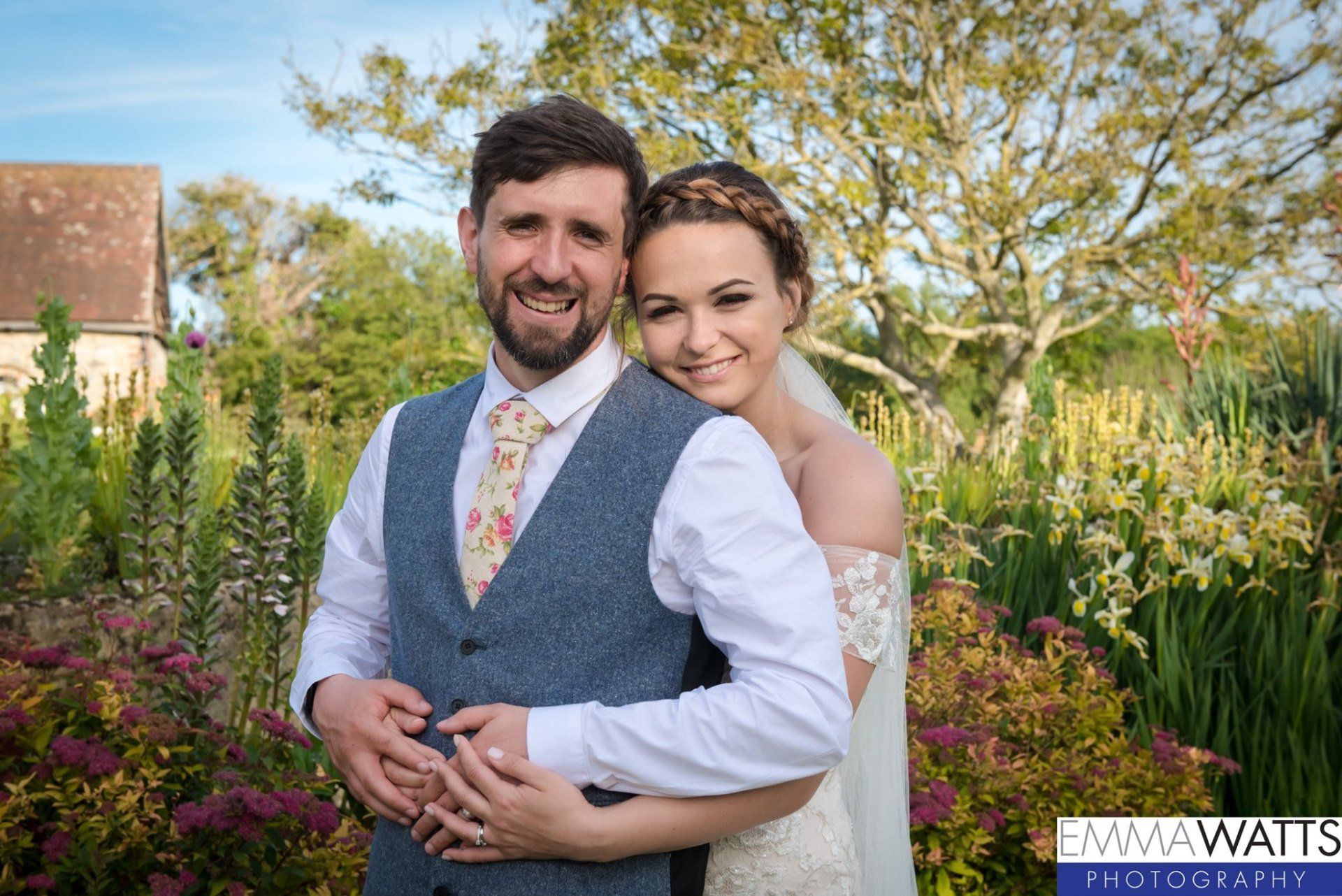 Newlyweds embrace outdoors; man hugs bride, both smile. Garden background, sunny.