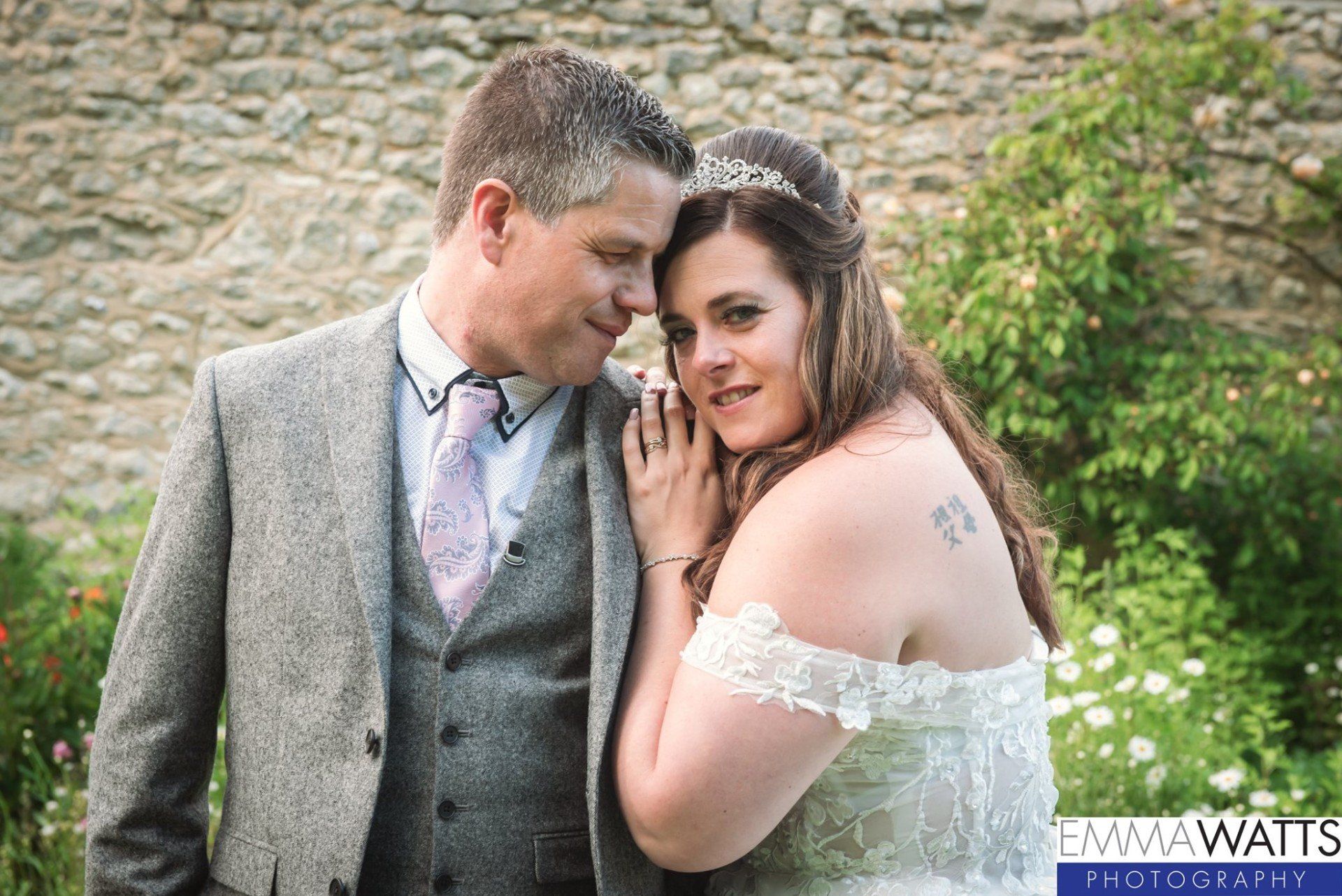 Couple in wedding attire embrace by a stone wall, smiling. Bride has off-the-shoulder dress, groom in tweed jacket.