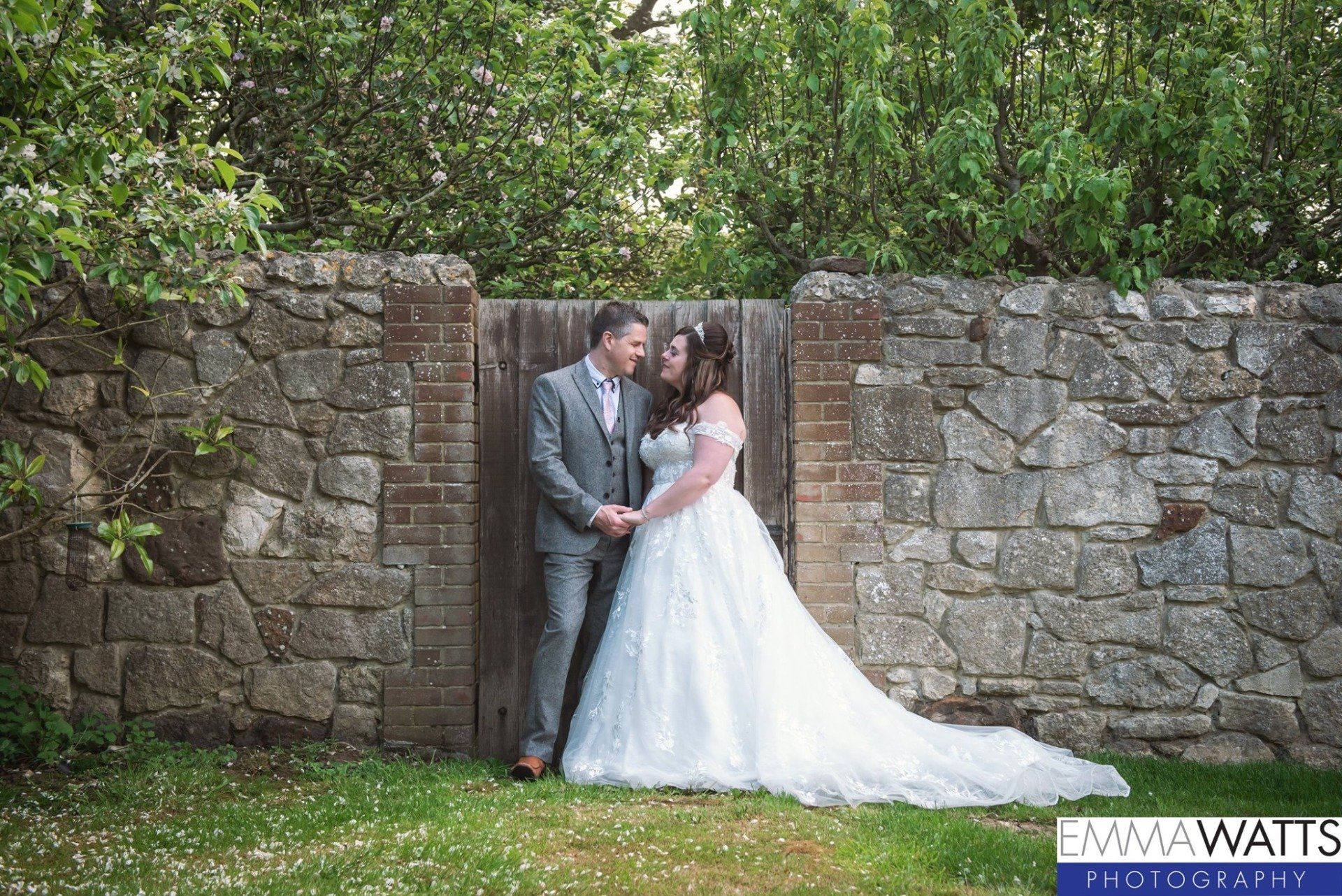 Couple in wedding attire hold hands, standing before a stone wall and wooden gate. They face each other, smiling.