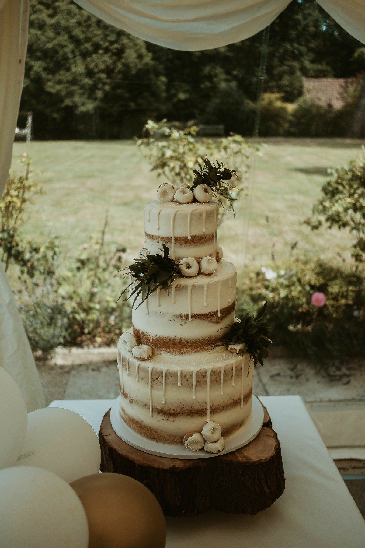 Four-tiered wedding cake with white frosting and donuts, on a wooden base, outdoors.