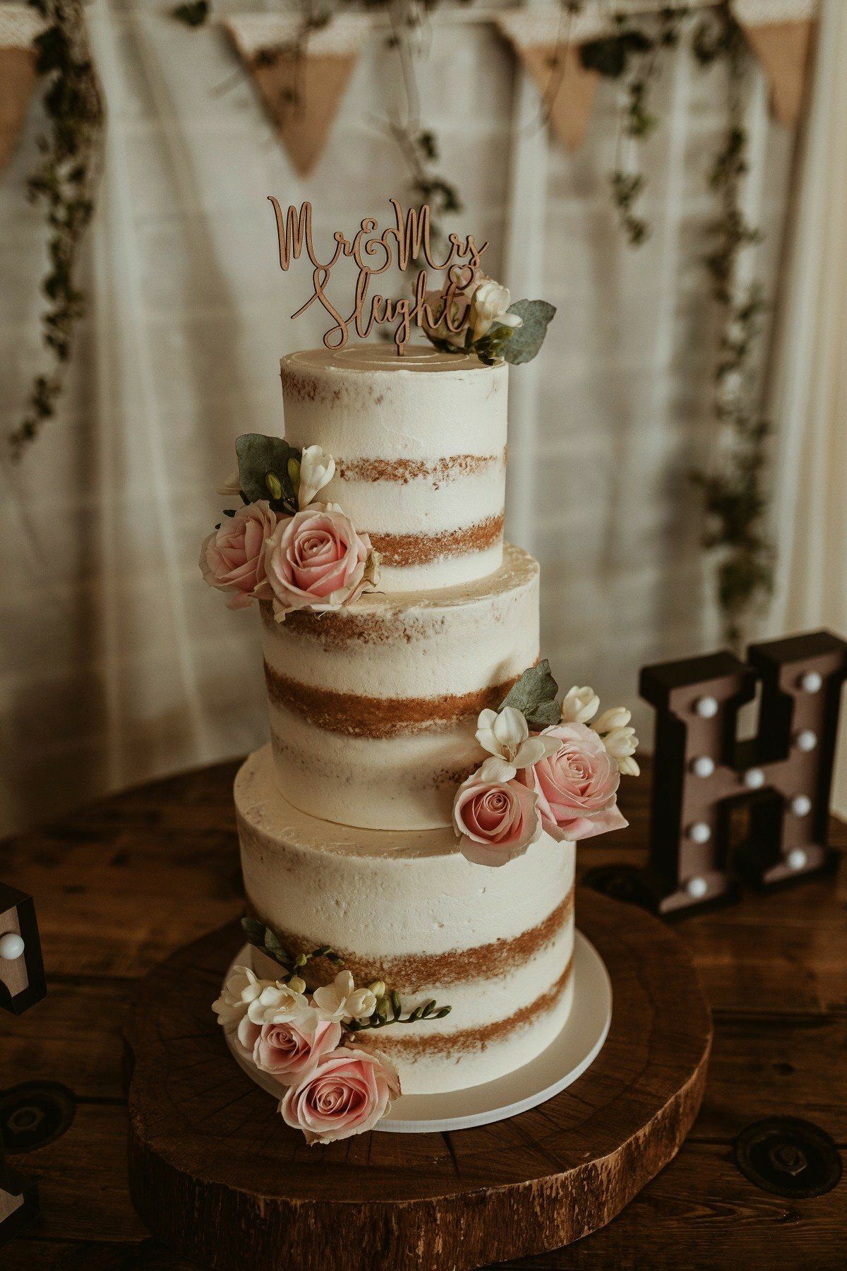 Four-tiered wedding cake, decorated with flowers and topper. Rustic wooden setting with draped greenery.
