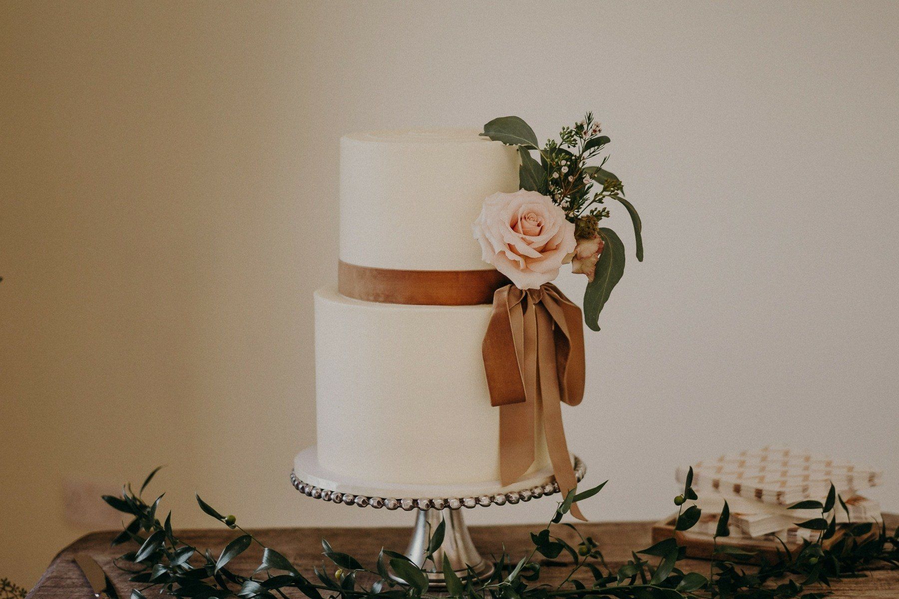 Two-tiered white frosted cake with a brown ribbon, rose, and greenery. On a pedestal.