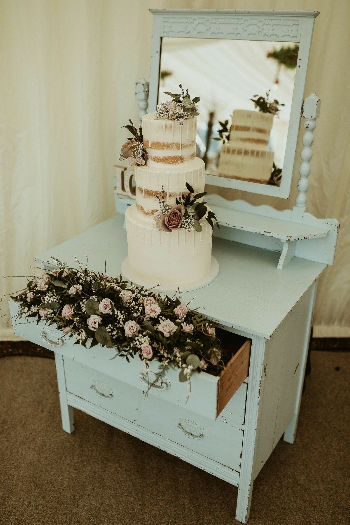 Three-tiered wedding cake on a light blue dresser with flowers, a mirror in the background.