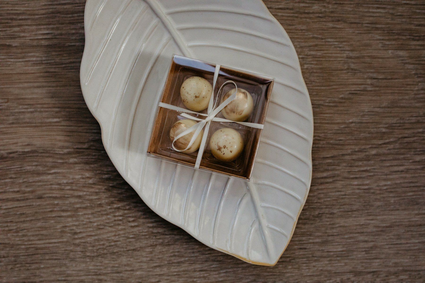 Four round chocolates in a clear box tied with string, placed on a leaf-shaped ceramic plate on wood.