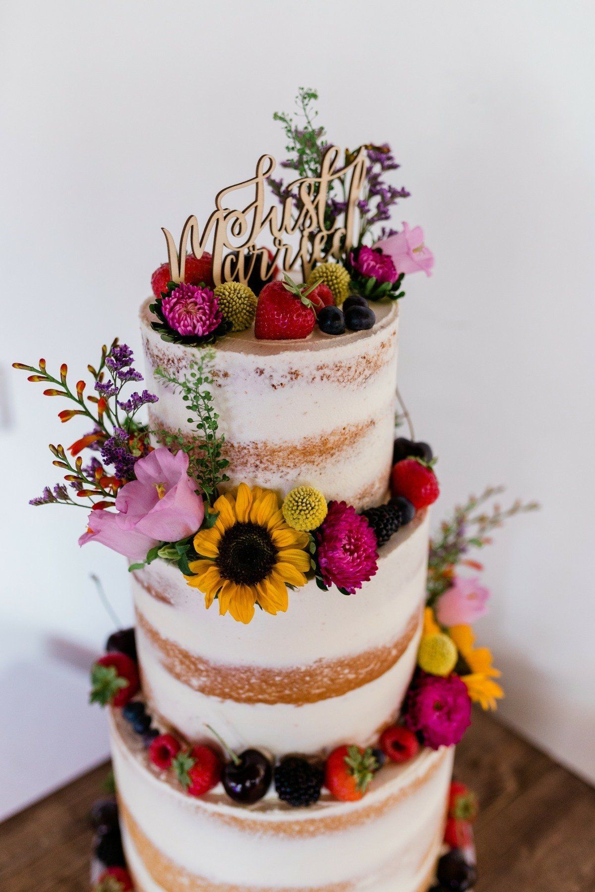 Three-tiered wedding cake with exposed layers, topped with fresh berries, flowers, and a wooden