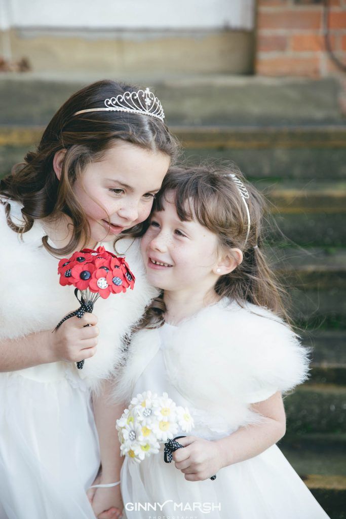 Two young girls in white dresses and fur shawls, holding bouquets, near a building.