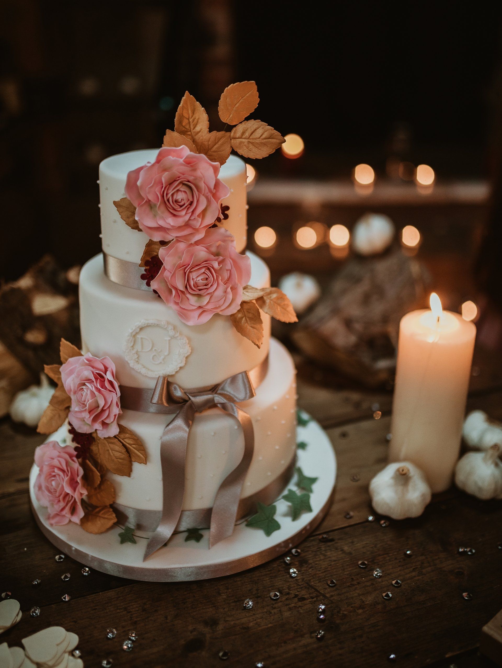 Three-tiered white cake with pink roses and gold leaves. Ribbon and candlelit setting.