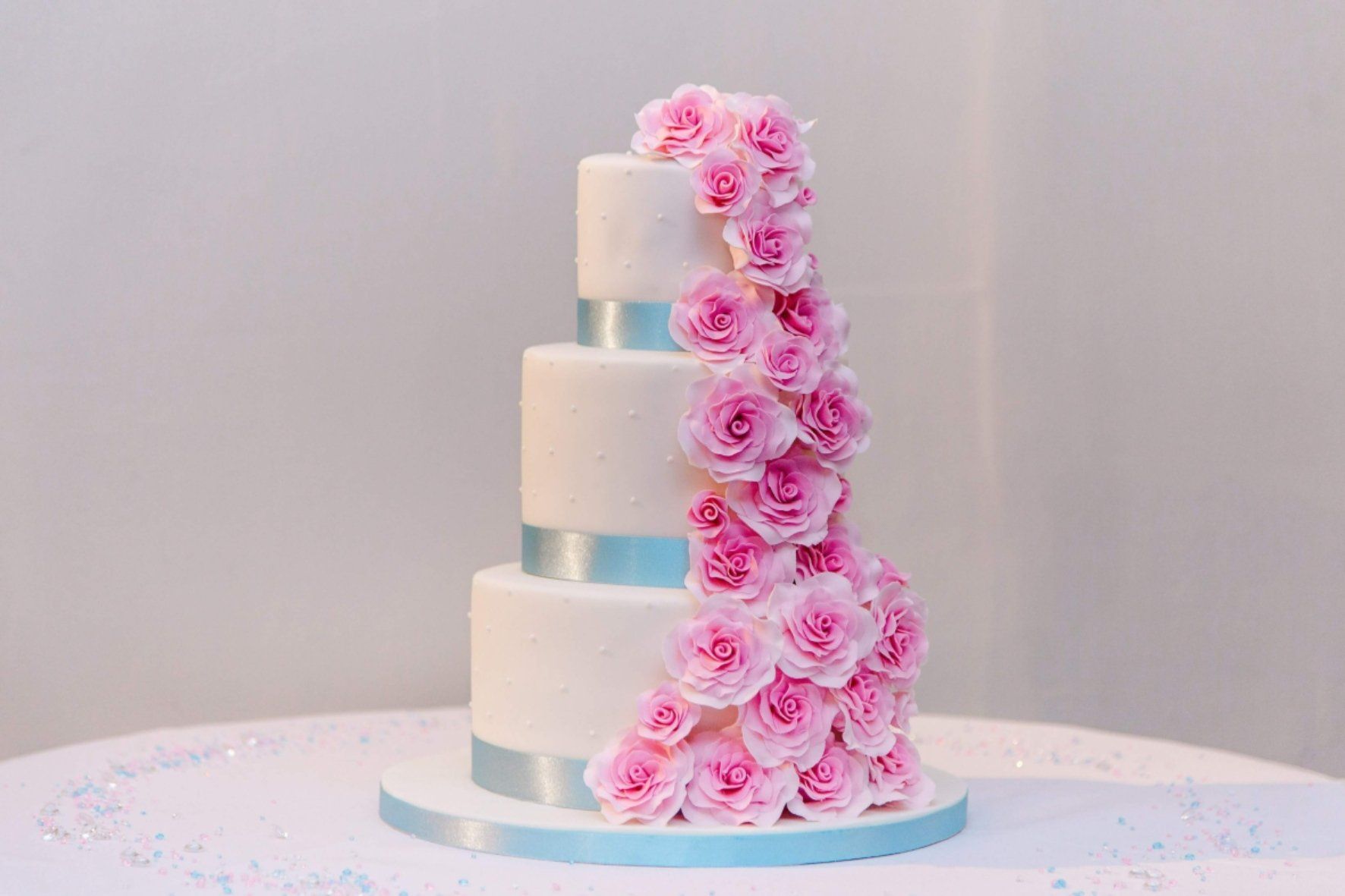 Three-tiered white cake with pink rose cascade and light blue ribbon on white surface.