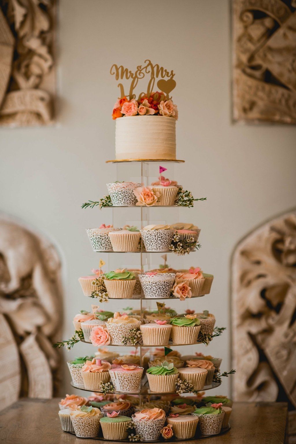 Wedding cake tower with cupcakes and topper. Beige, peach, and green decorations, inside setting.