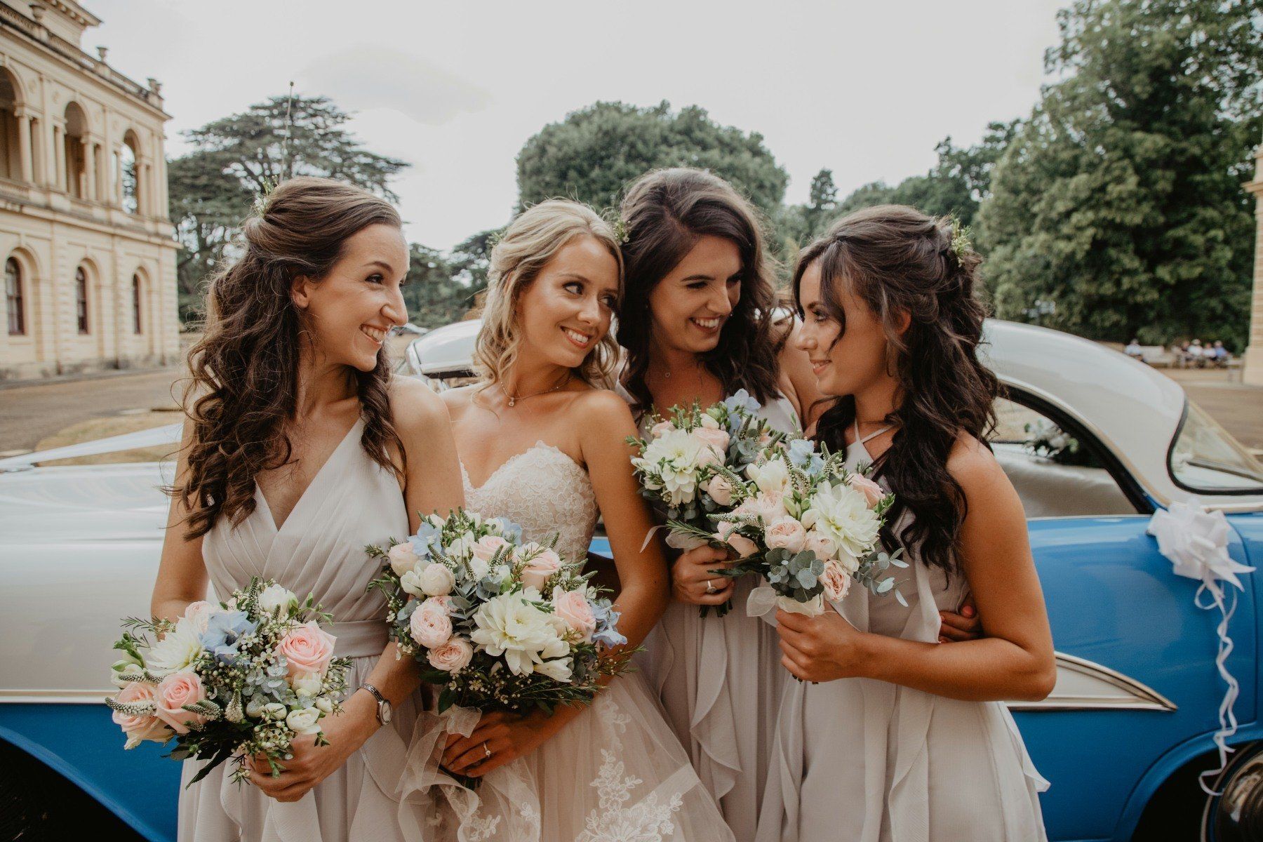 Bridesmaids with bouquets smile near a vintage blue car in front of a building.