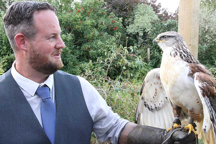 Man in vest and tie, holding a hawk with brown and white feathers in an outdoor setting.