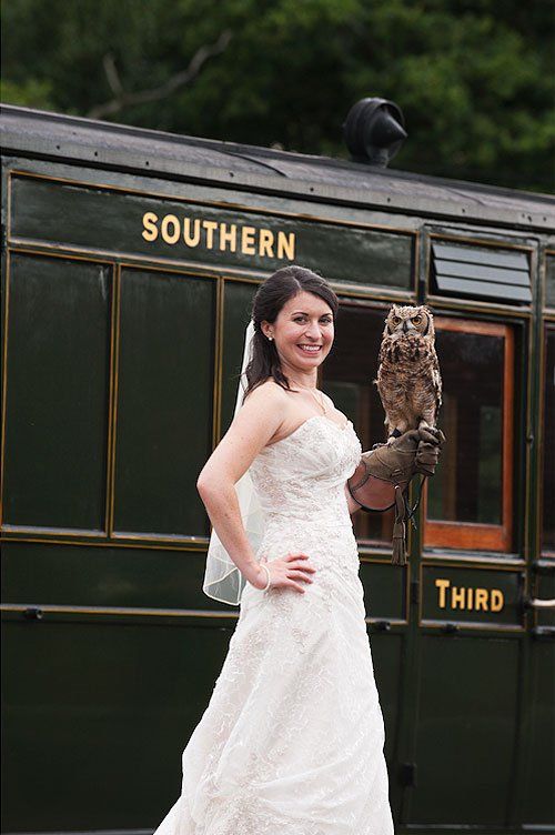 Bride in white dress holds an owl next to a vintage Southern train car.