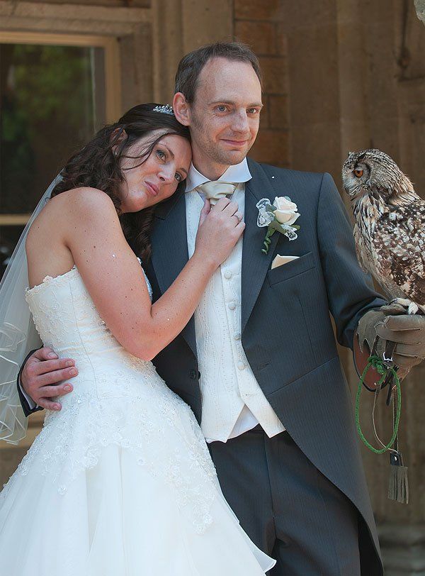 Bride and groom pose with an owl on their wedding day; woman leans on man's shoulder.