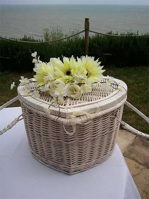 Wicker heart-shaped basket with flowers atop, set on white tablecloth outdoors with ocean view.