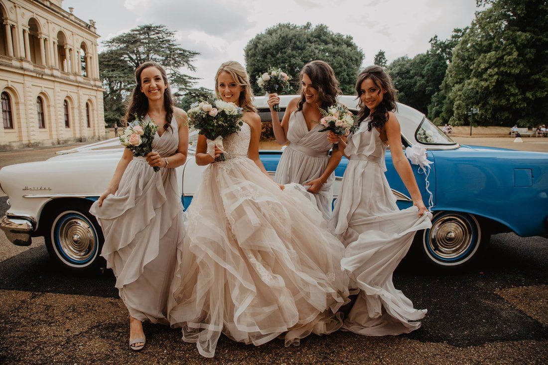 Bride and bridesmaids in flowing gowns, holding bouquets, near a blue vintage car in front of a grand building.