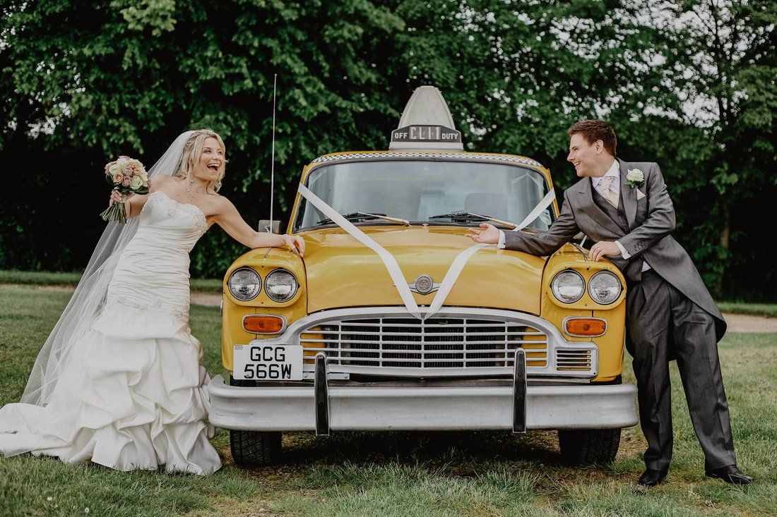 Bride and groom lean on a yellow taxi decorated with ribbons, smiling and laughing outdoors.