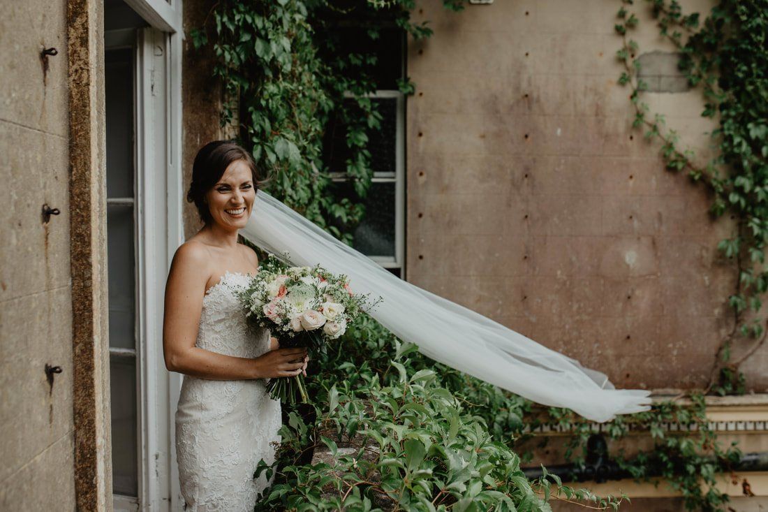 Bride holding bouquet, veil blowing in the wind, standing by a doorway with ivy growing on a building.
