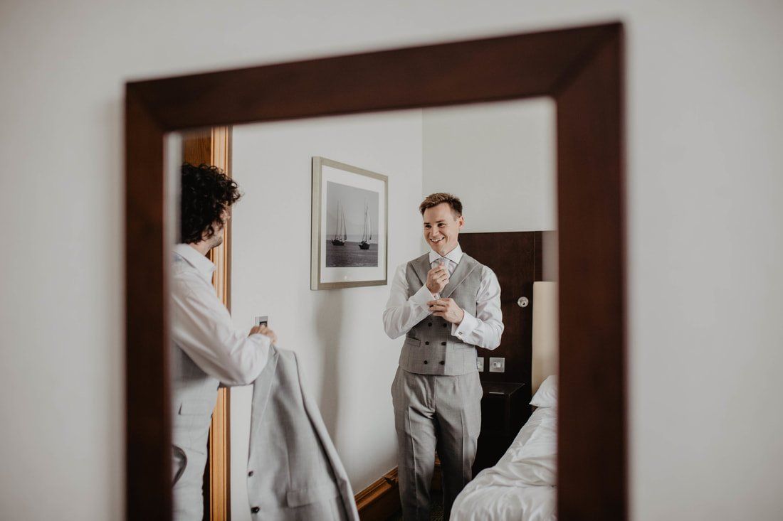 Man in gray suit adjusts tie in mirror, another man holds a jacket.