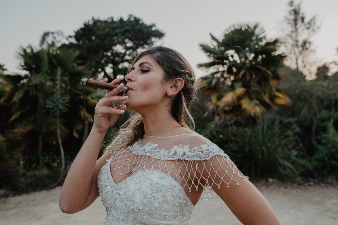 Bride in a wedding dress smoking a cigar outdoors, with trees in the background.