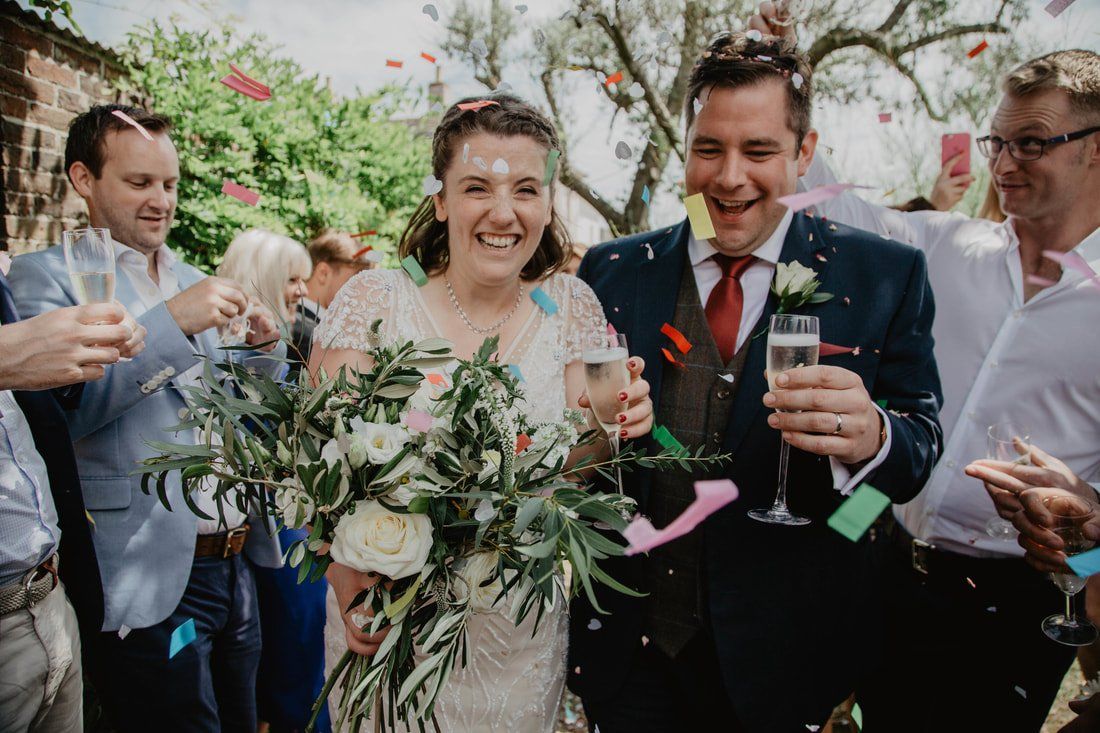 Newlyweds smiling, showered in confetti, holding champagne, and bouquets outside.