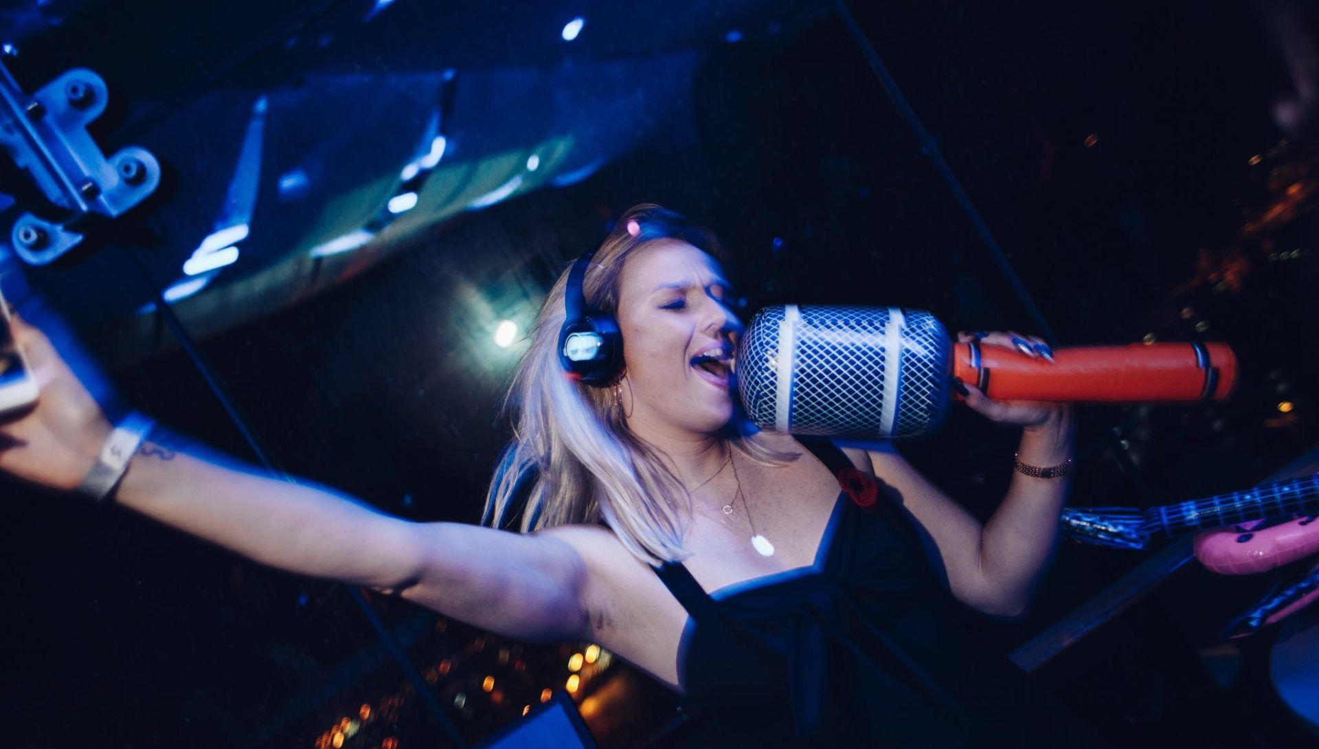 Woman singing into a large, toy-like microphone, wearing headphones, in a dark, blue-lit setting.