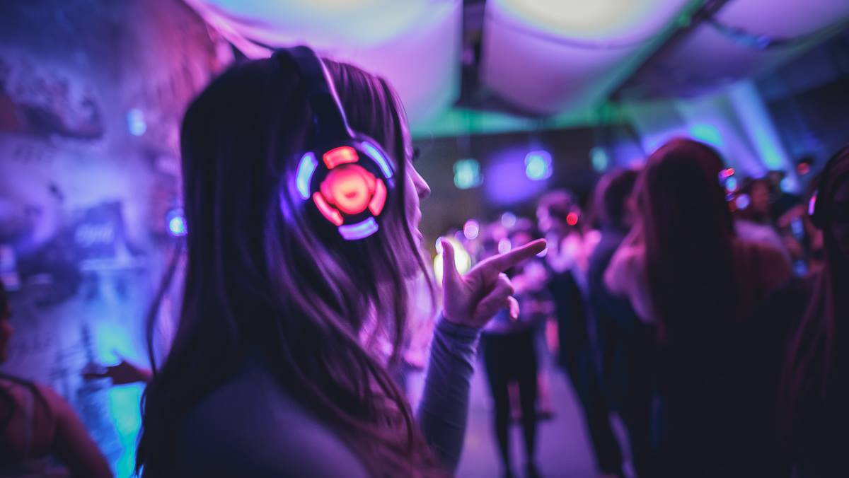 Woman with glowing headphones dancing at a silent disco, bathed in purple and blue lights.