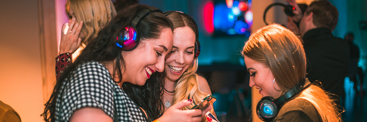 Three women smile and laugh while looking at a phone in a dimly lit setting with headphones on.
