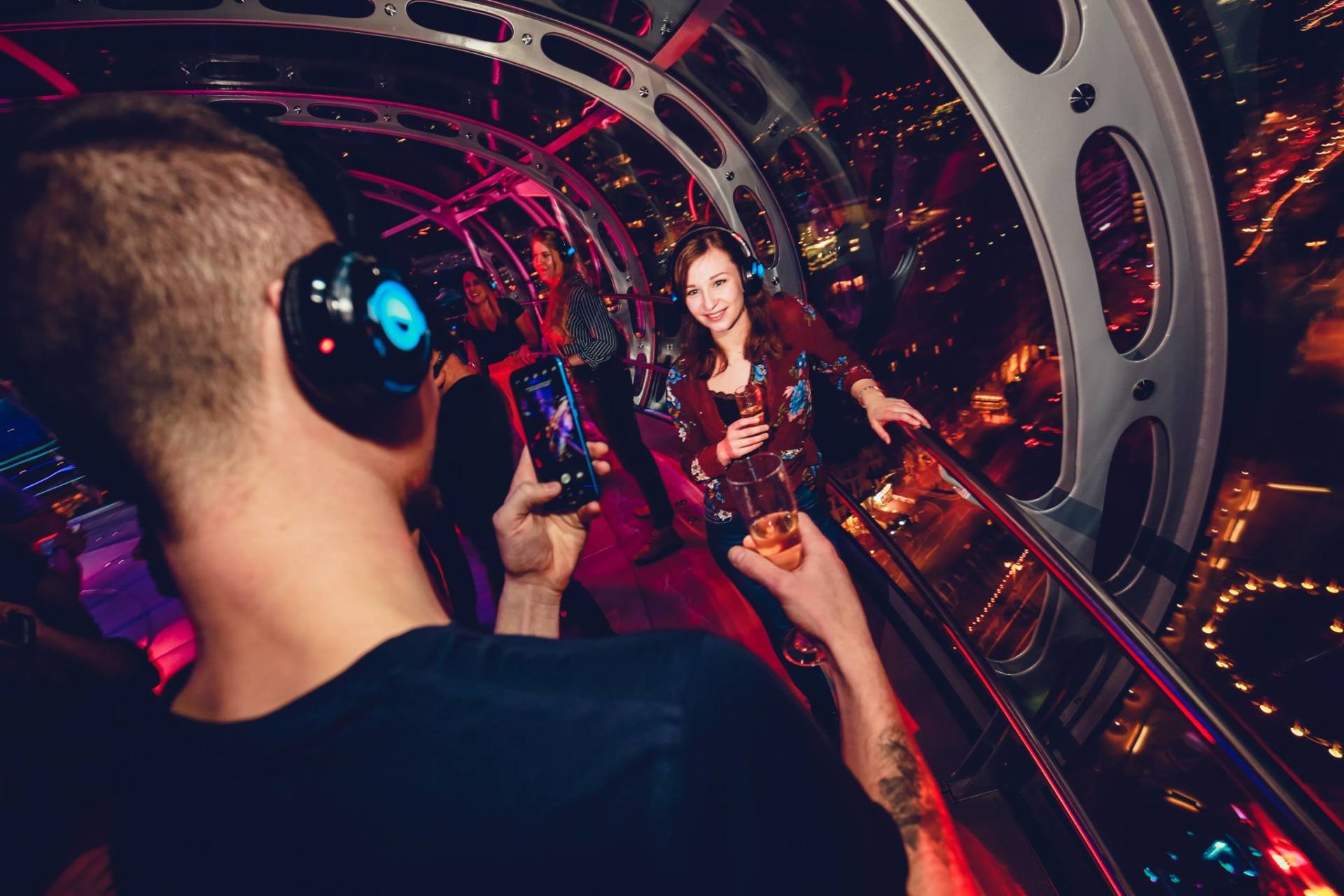 People in a silent disco on a high, enclosed walkway with city lights. One person holding a phone, another smiling.