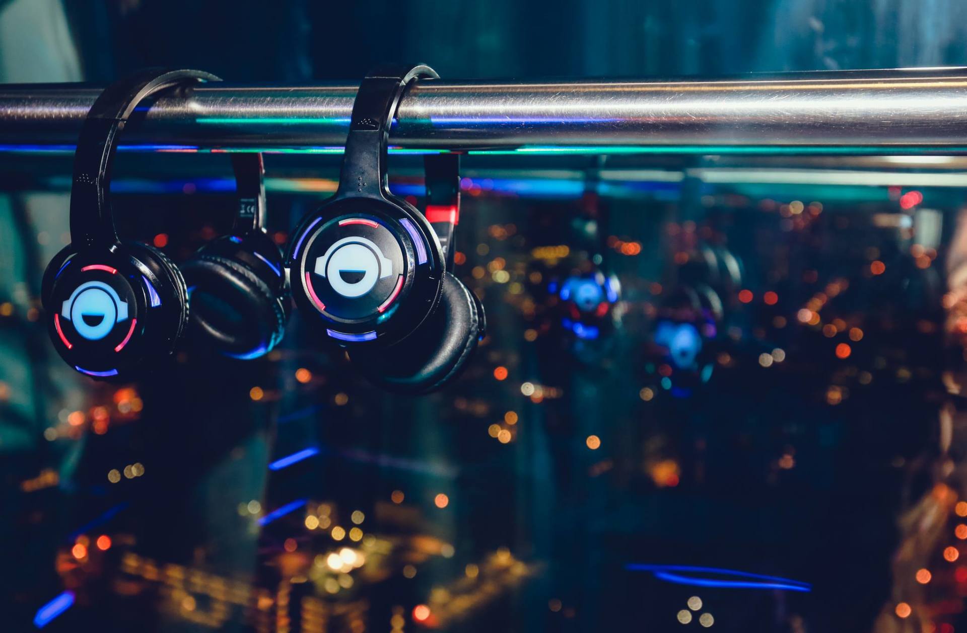 Headphones with glowing blue and red lights hanging from a metal bar with a blurry city backdrop.