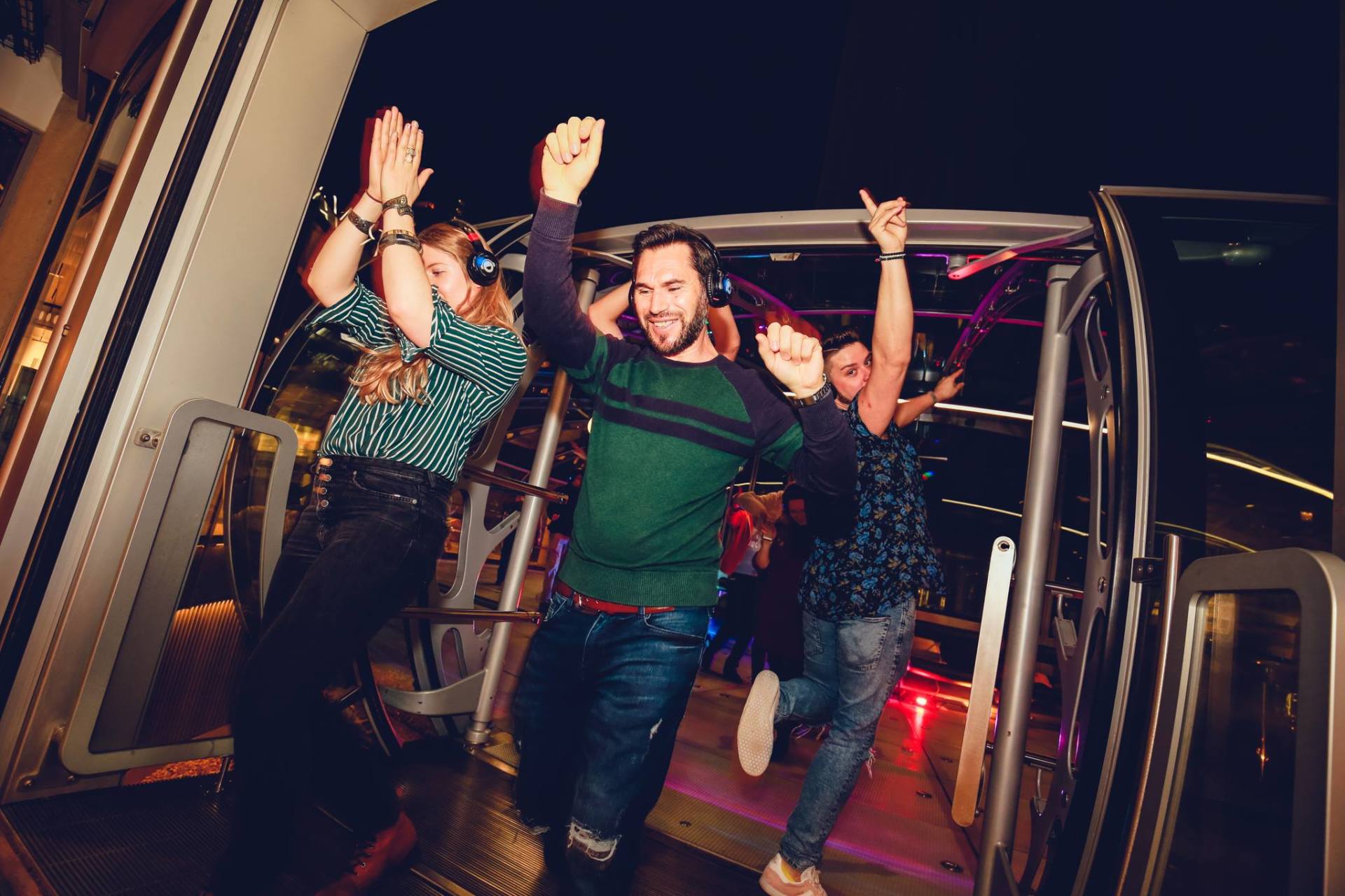 People dancing on a bus at night, wearing headphones, arms raised, lit by colorful lights.