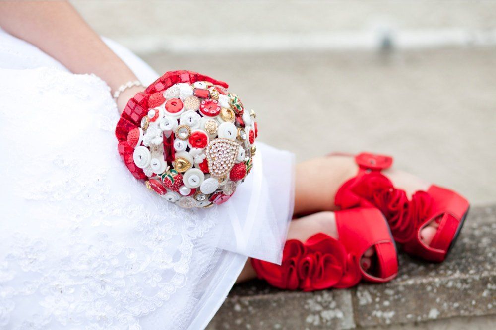 Bride in white dress, red shoes, holding a bouquet of red and beige buttons, sitting outside.