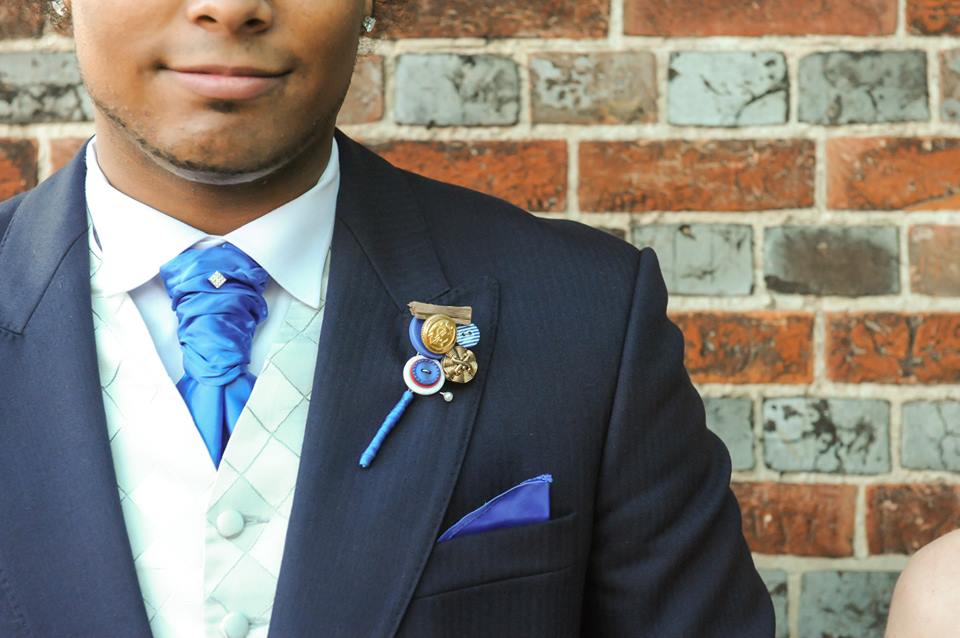 Man in a blue suit with a blue tie, white vest, and a boutonniere. Leaning against a brick wall.