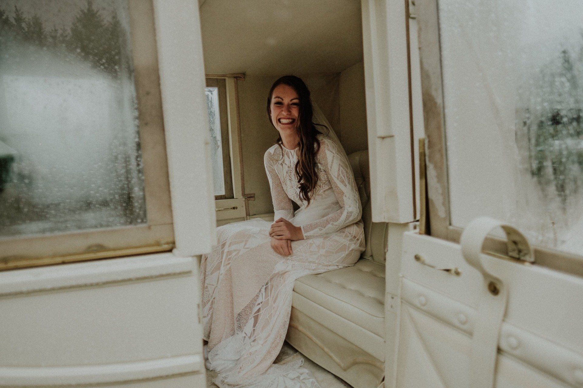Smiling woman in a wedding dress inside a white carriage, looking out a window.