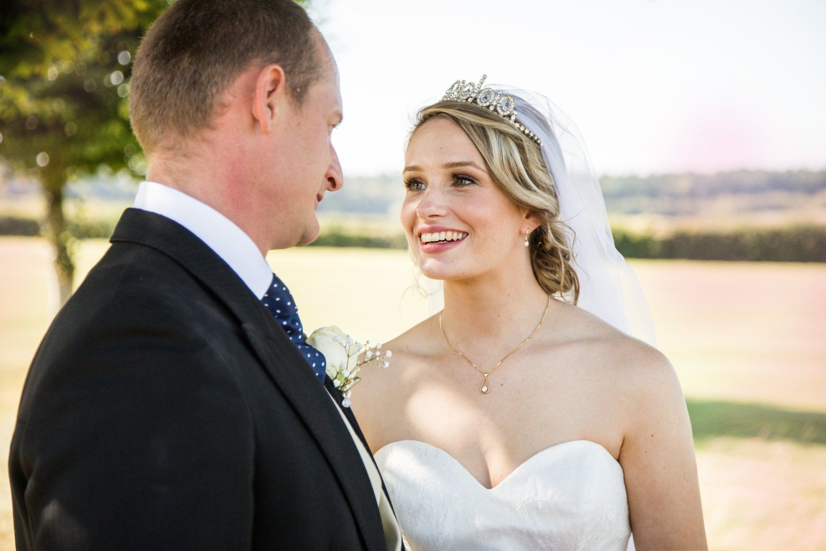 Bride in white strapless dress smiles at groom in suit, outdoor setting, fields visible.