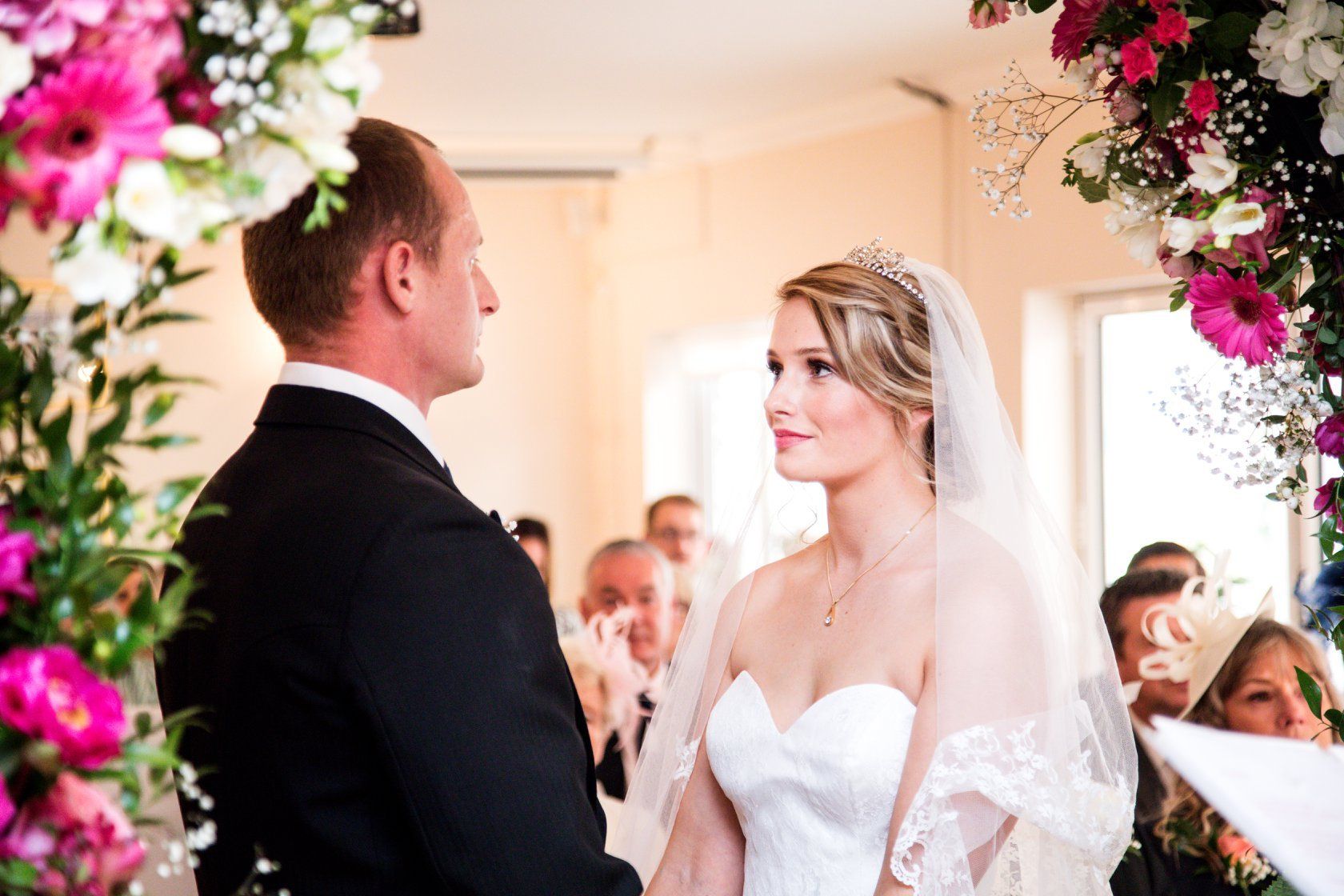 Bride and groom at wedding ceremony, exchanging vows, surrounded by floral arch, guests in background.