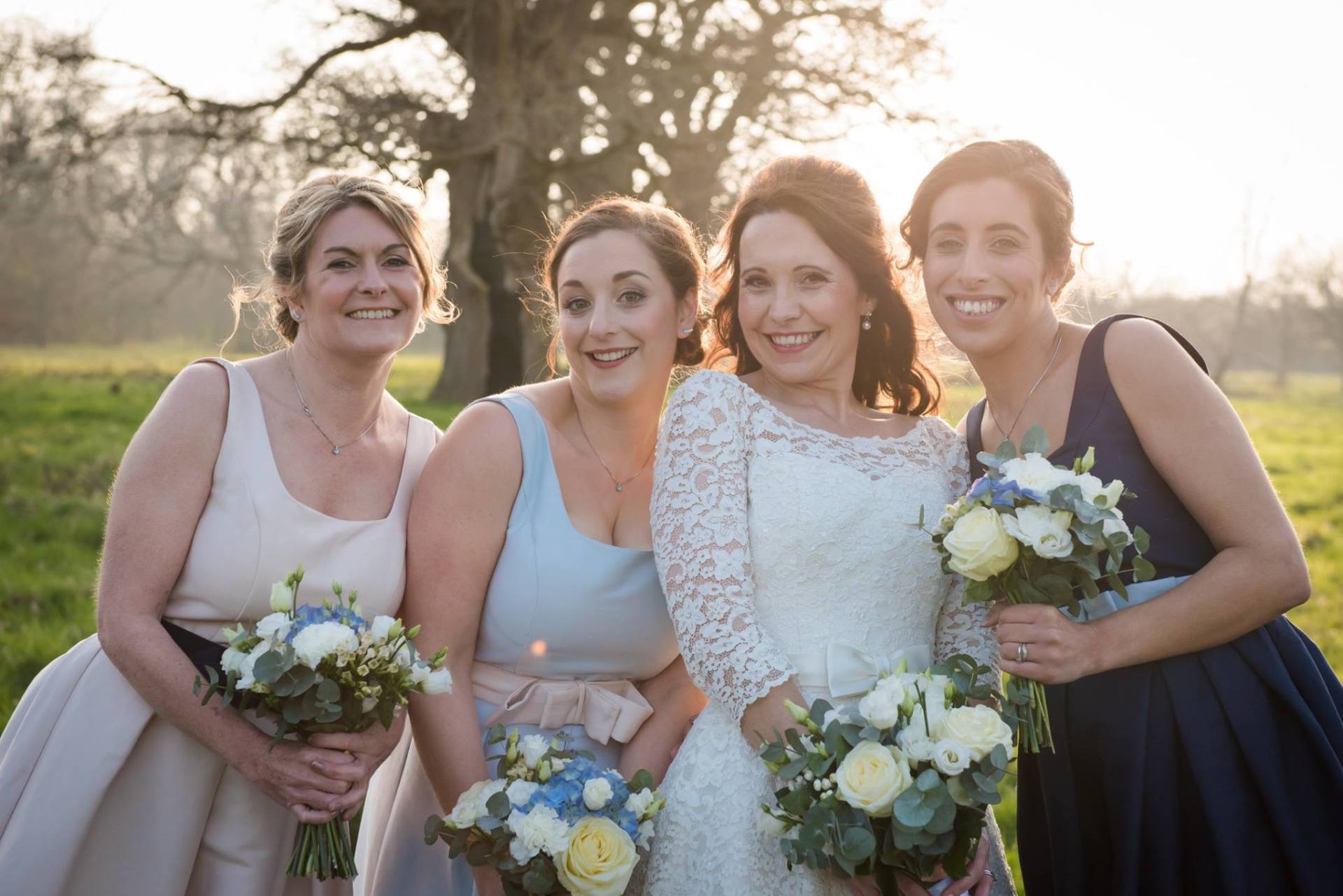 Bride and bridesmaids smiling outdoors with bouquets, soft light.