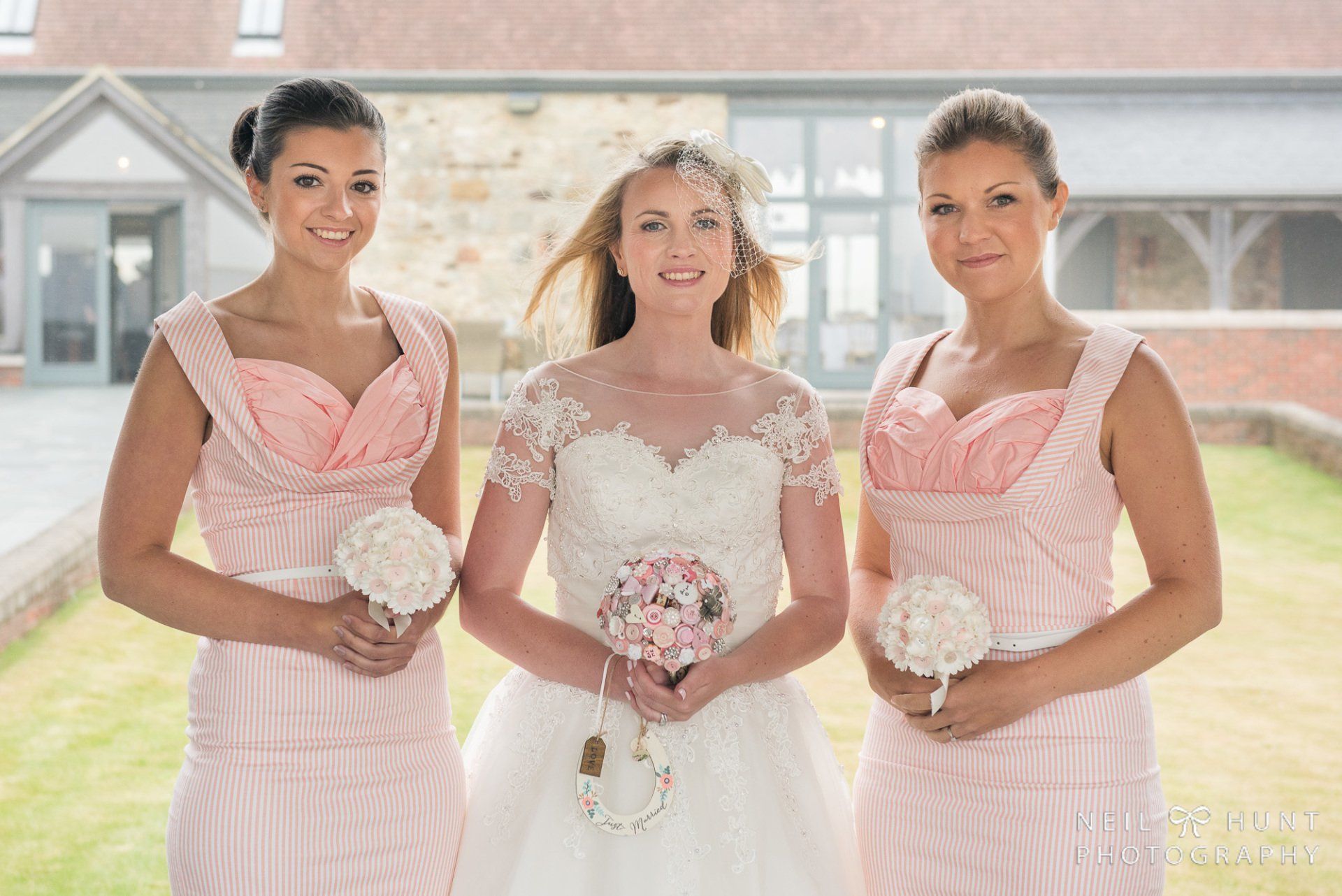 Bride with two bridesmaids holding bouquets, all smiling. Bride in white, bridesmaids in pink, outdoors.
