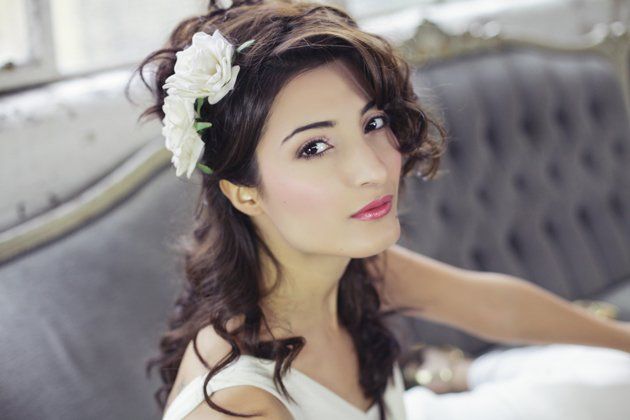 Woman with wavy brown hair, flower hairpiece, light makeup, on a grey couch.