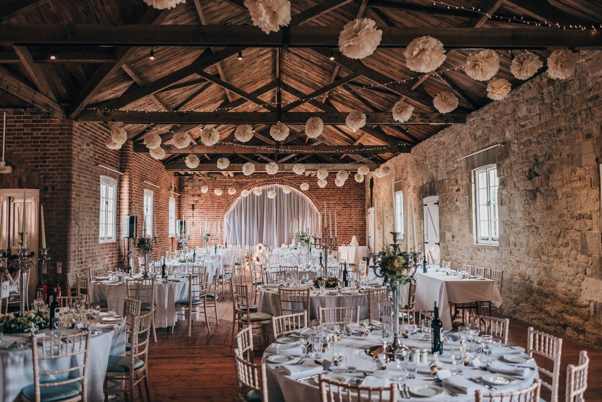 Wedding reception hall, round tables set with white linens and tableware, decorated with floral centerpieces and paper pom-poms.
