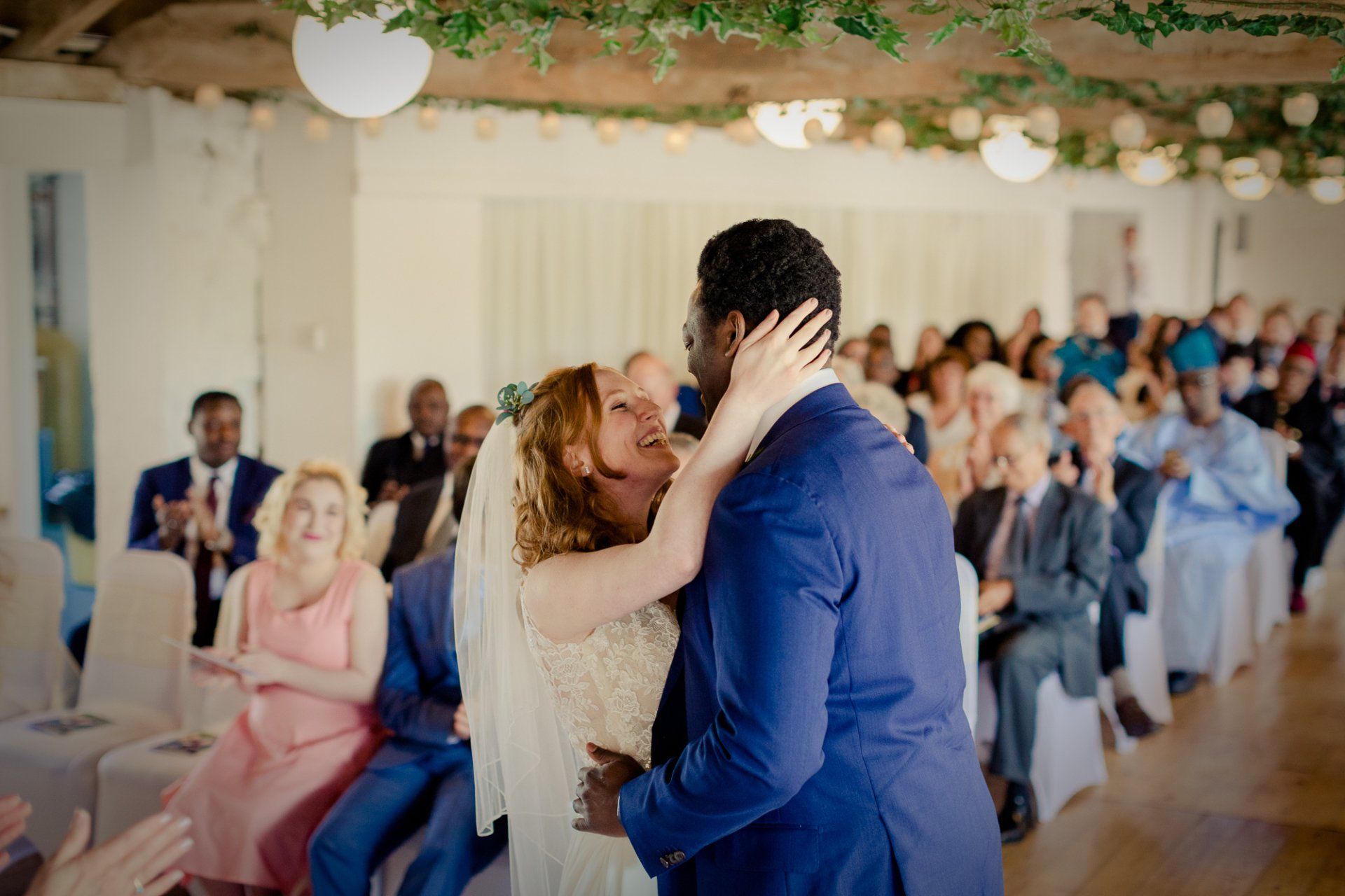 Newlyweds embrace at the altar, guests clap. White building, floral decor overhead.