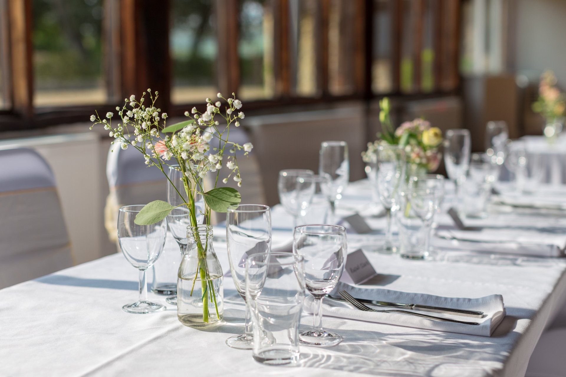 Table set for event with white tablecloth, glassware, and small floral arrangements.