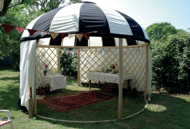 A black and white striped gazebo with tables, rug, and draped curtains, set in a sunny garden.