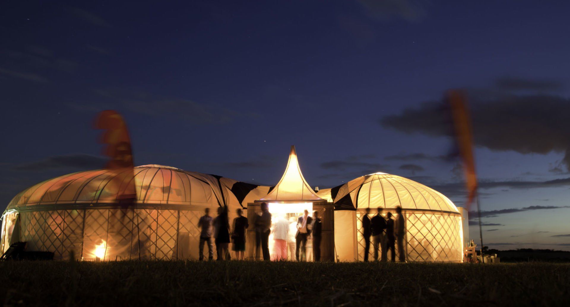 Yurts at dusk, illuminated from within. People stand in doorways; orange smoke rises.