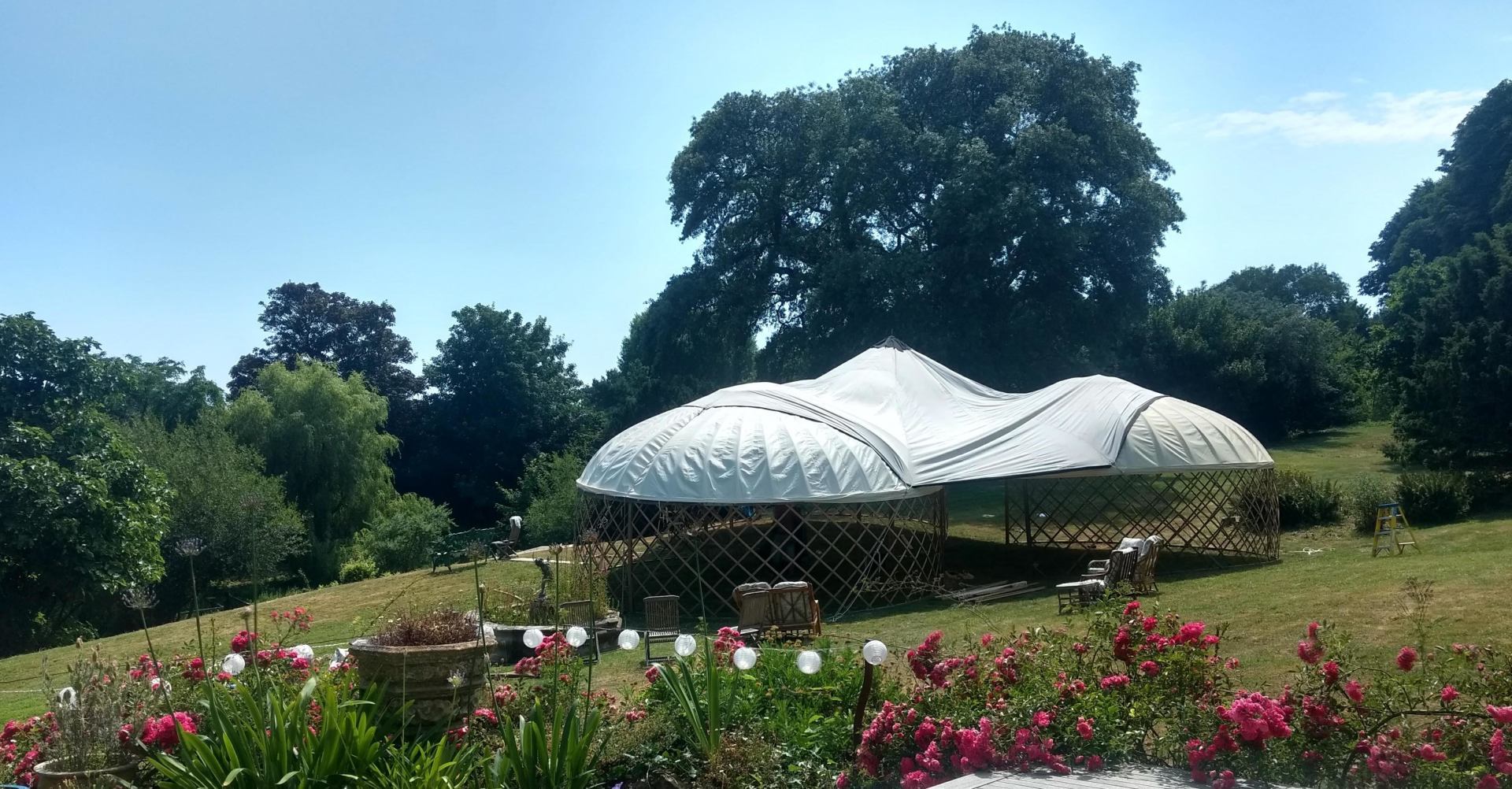 A white-roofed tent structure in a grassy garden, surrounded by trees and flowers under a blue sky.