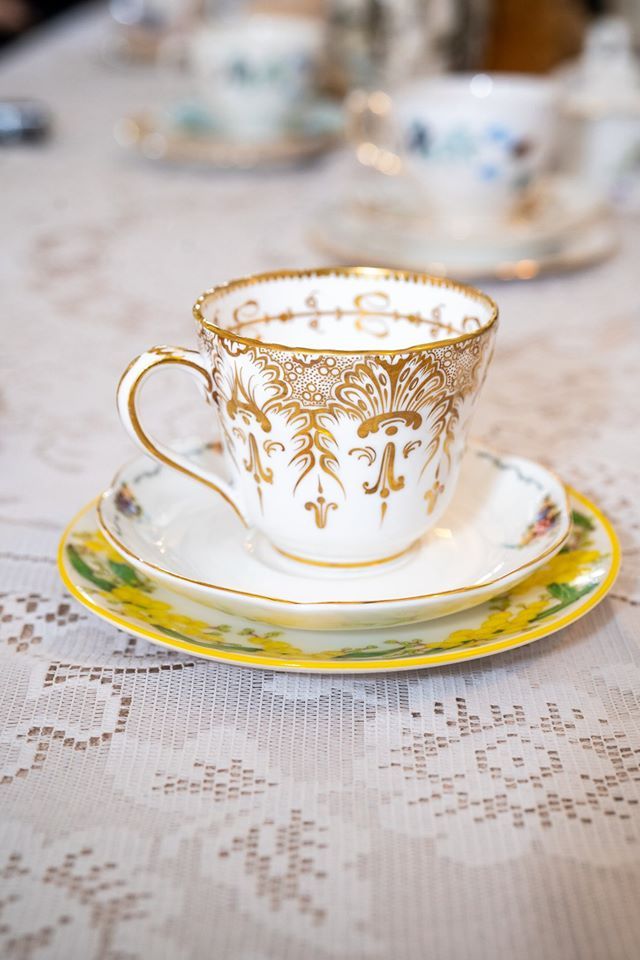 Elegant white teacup with gold filigree, sits on a saucer with a yellow rim, on a lace tablecloth.